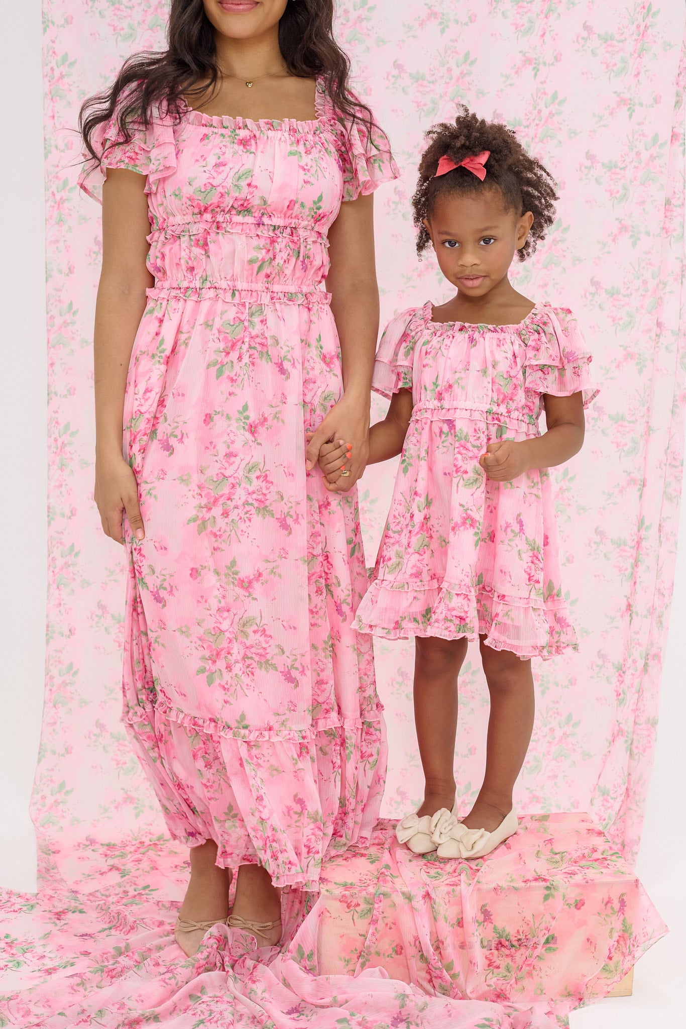 A woman and a young girl hold hands in matching Ivy City Co's Juliana Dress in Rose Trance. The girl stands on a box surrounded by the same pink floral fabric—these maternity-friendly dresses are perfect for moms.