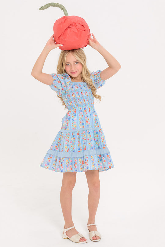 young girl in a blue floral dress holding a red paper cherry above her head standing against a white background