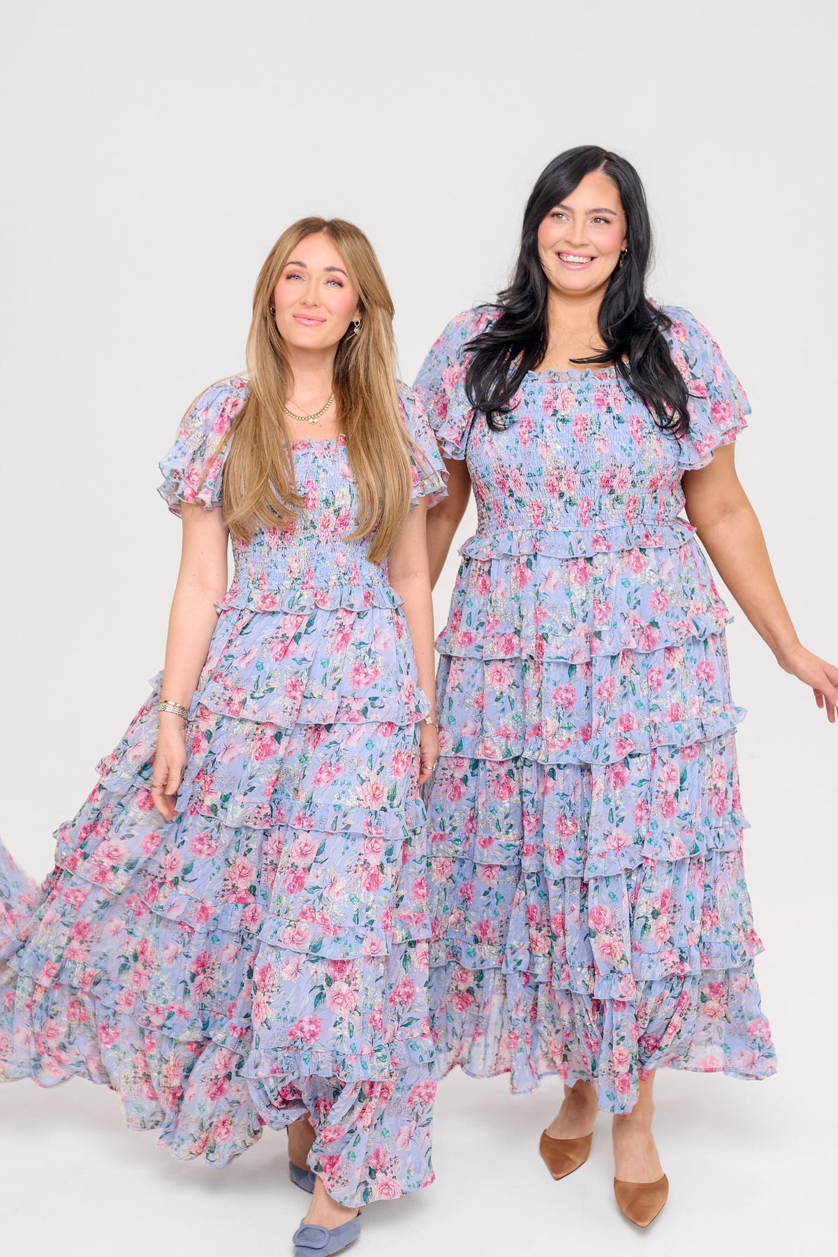 Two women stand smiling in matching Ivy City Co Winnie Dresses in Peony Blossoms—maternity-friendly, tiered floral maxi dresses with pink patterns and short puff sleeves—against a plain white background.