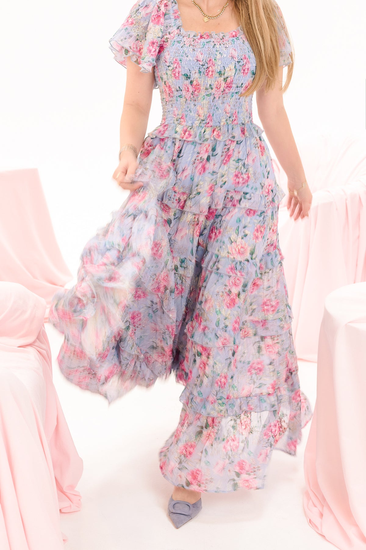 A woman in the Ivy City Co Winnie Dress in Peony Blossoms, a floral short-sleeve maxi, walks among pale pink-draped tables. Only her lower face is visible and she wears lavender pointed-toe shoes. The look is maternity friendly.