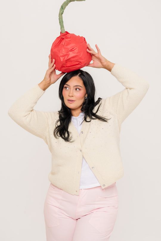 A woman with long dark hair wears the Ivy City Co Remy Sweater in Cream and holds a large red paper-mâché cherry with a green stem on her head, standing against a plain light background.
