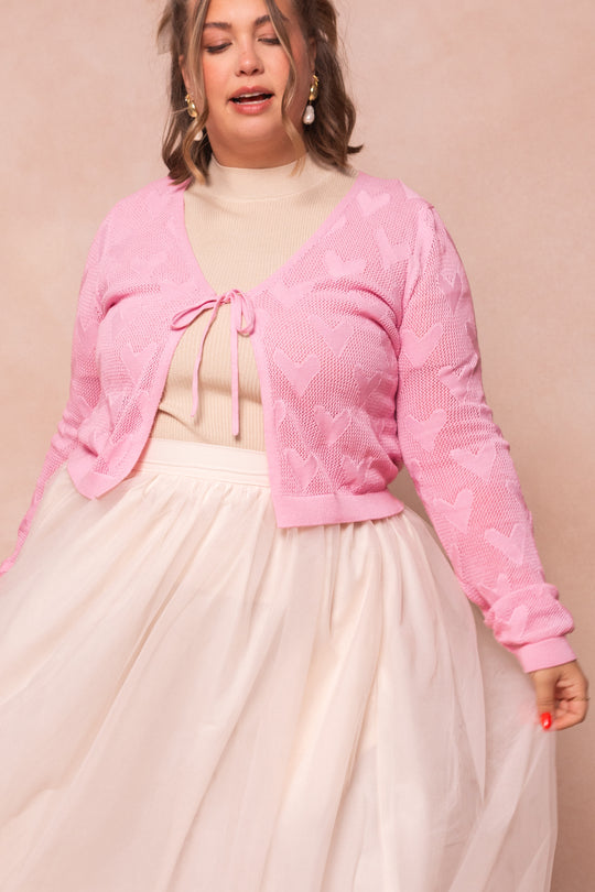 A woman models the Ivy City Co With Love Sweater Cardigan, featuring a light pink heart pattern, layered over a cream turtleneck and tulle skirt, posed against a soft pink background.