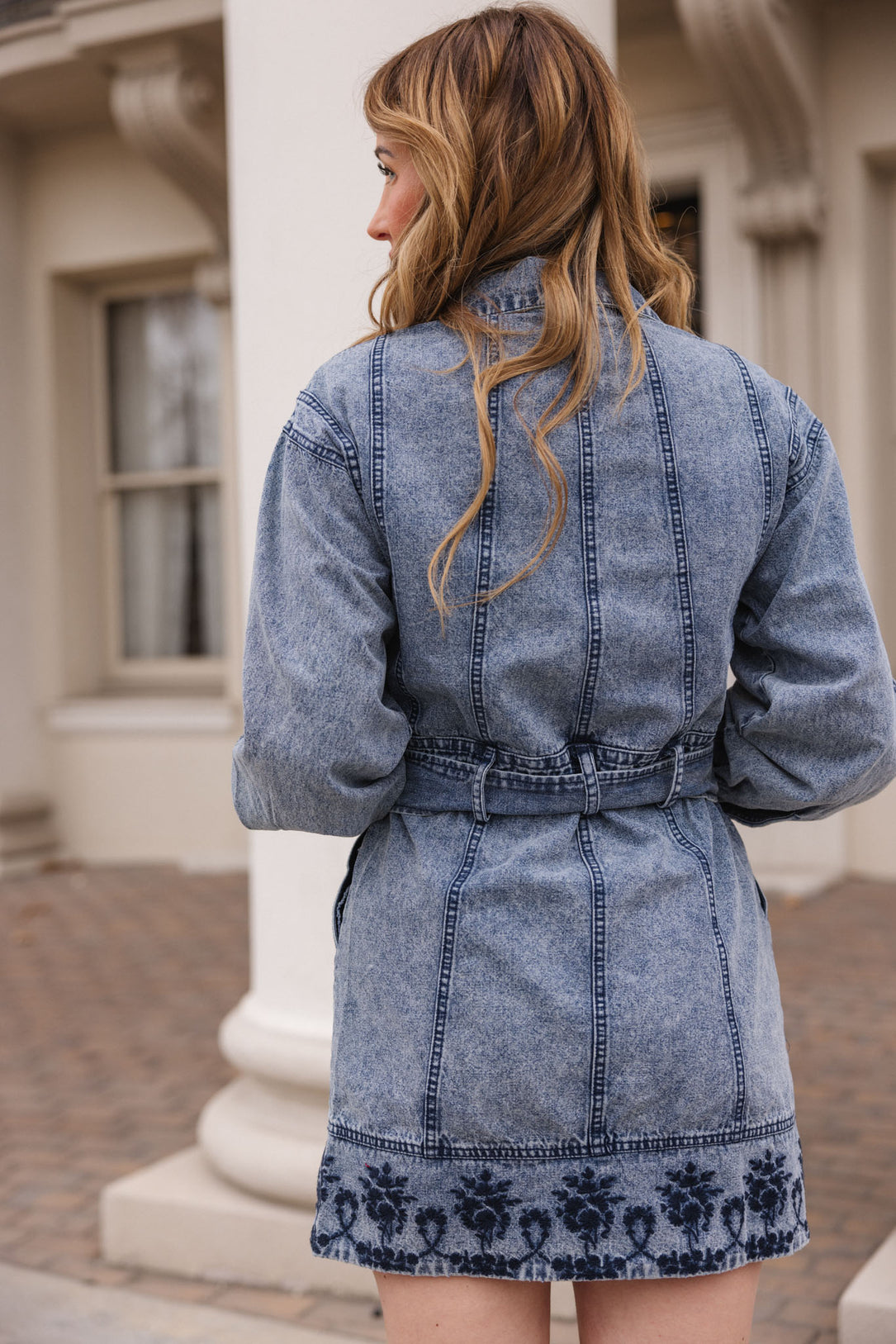 A woman with long, wavy hair stands outside facing away, wearing the Ivy City Co Elodie Dress in Light Wash Denim. The dress features a structured silhouette and dark decorative stitching, set against a building with white columns and windows.