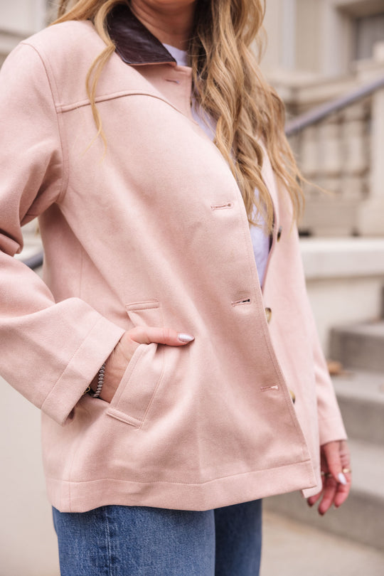A woman with long, wavy blonde hair wears the Ivy City Co Daphne Coat in Light Pink, paired with blue jeans and a white shirt. She stands outdoors near concrete steps and a railing, her left hand in her pocket.