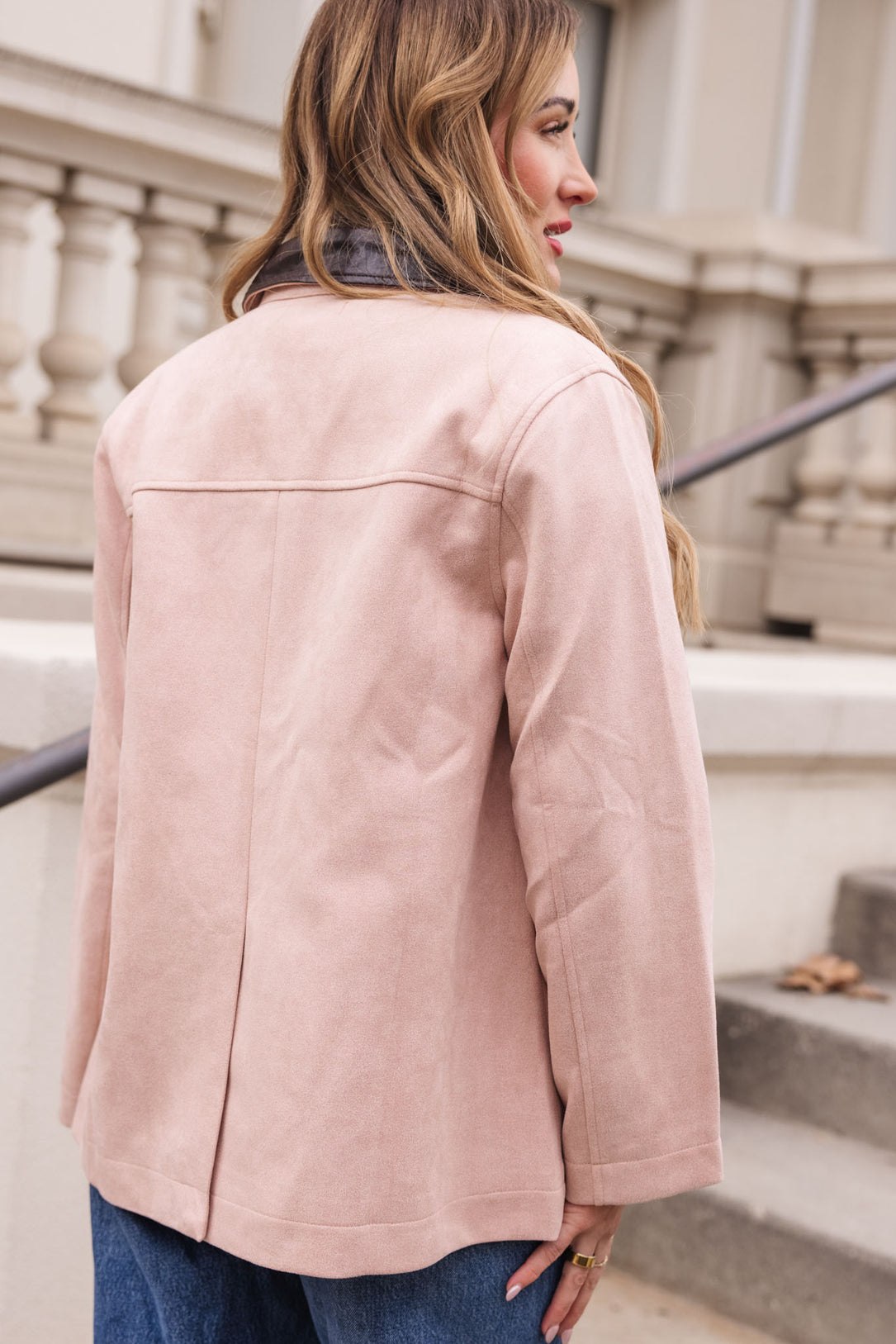 A woman with long, wavy hair stands outdoors facing away, wearing the Ivy City Co Daphne Coat in Light Pink and blue jeans near a staircase with a decorative railing and a neutral building in the background.