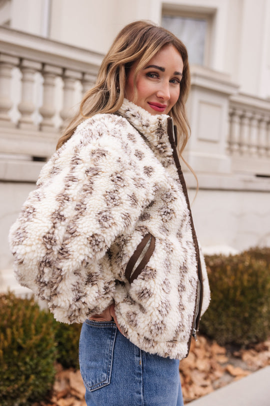 A woman with long, wavy hair stands outdoors in front of a building with columns, smiling at the camera. She wears Ivy City Co's Theodore Jacket in Tan & White and blue jeans, turned slightly sideways.
