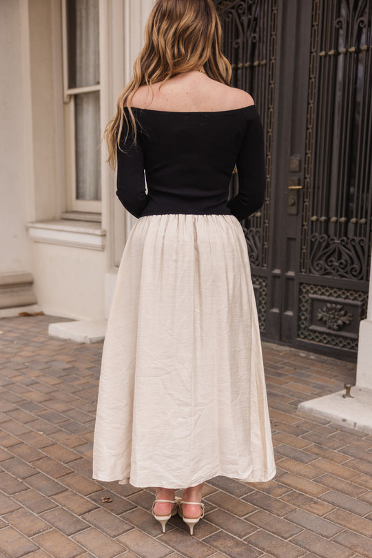 A woman with long, wavy hair stands on a brick walkway facing ornate dark doors and tall windows, wearing the Ivy City Co Samara Dress in Black & White paired with pointed-toe heels.