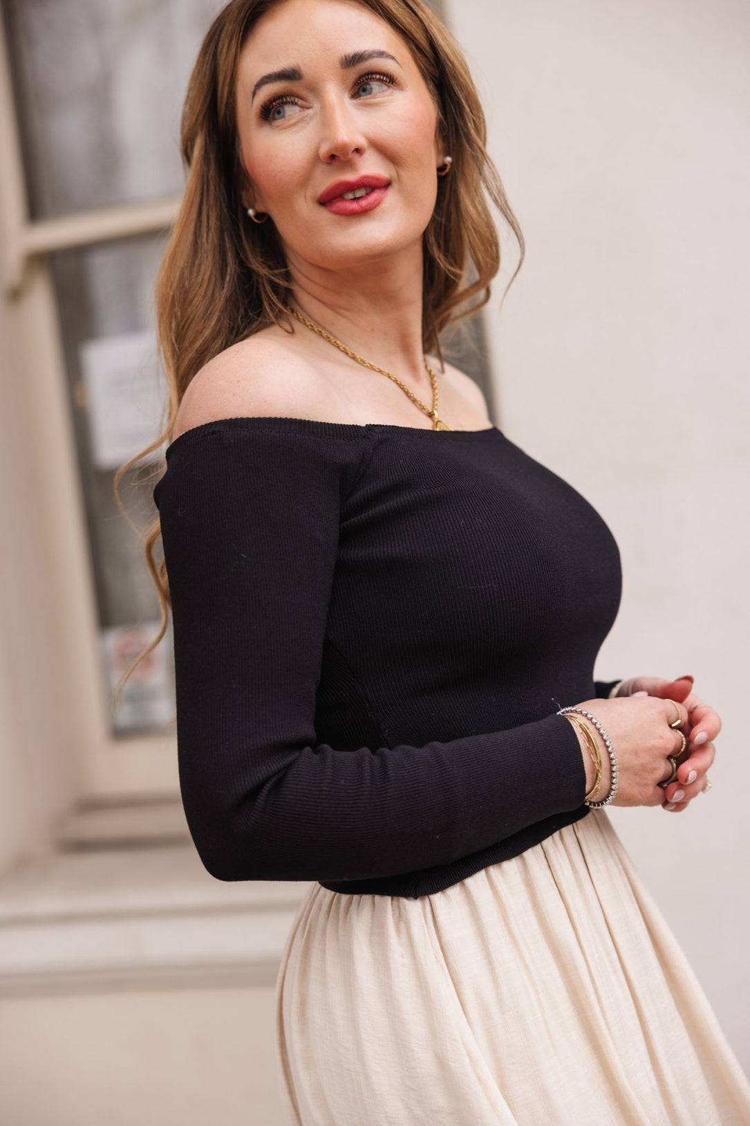 A woman with long, wavy hair stands outdoors wearing the Ivy City Co Samara Dress in Black & White, accessorized with gold jewelry and bracelets, paired with a cream-colored skirt as she looks off to the side and smiles.