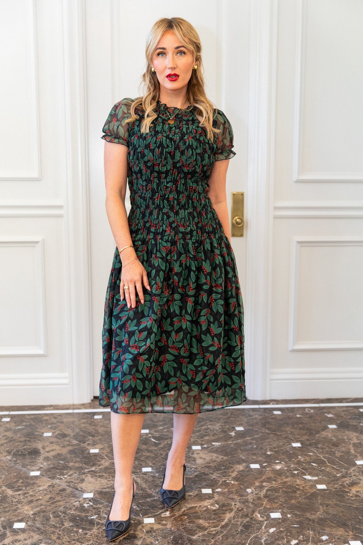 A woman wearing an Ivy City Co Hayden Dress in Berries with a smocked bodice, green leaf and red berry print, paired with black pumps.