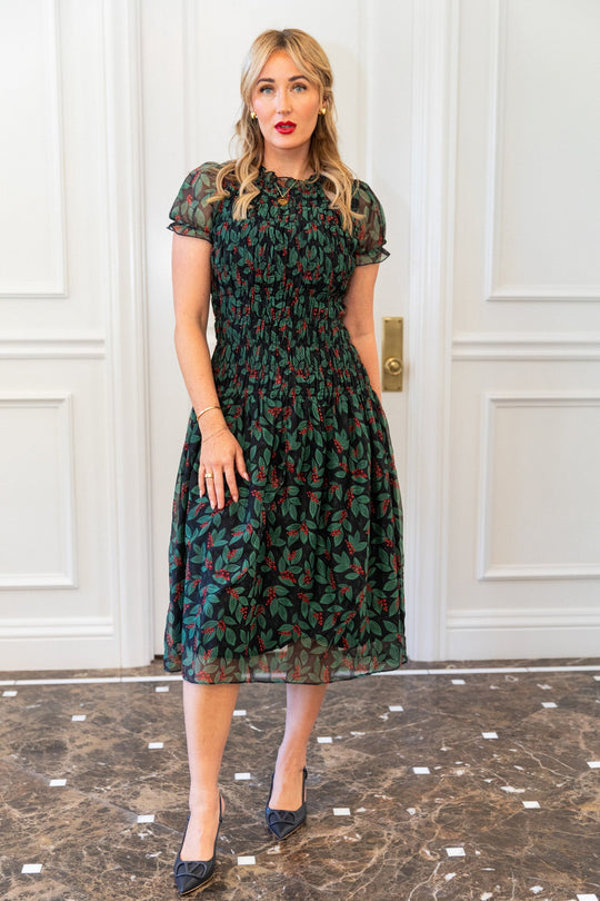 A woman wearing an Ivy City Co Hayden Dress in Berries with a smocked bodice, green leaf and red berry print, paired with black pumps.