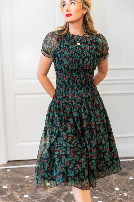 A woman wears an Ivy City Co Hayden Dress in Berries, a knee-length, short-sleeved dress with a smocked bodice, a black background, and a green leaf and red berry print.