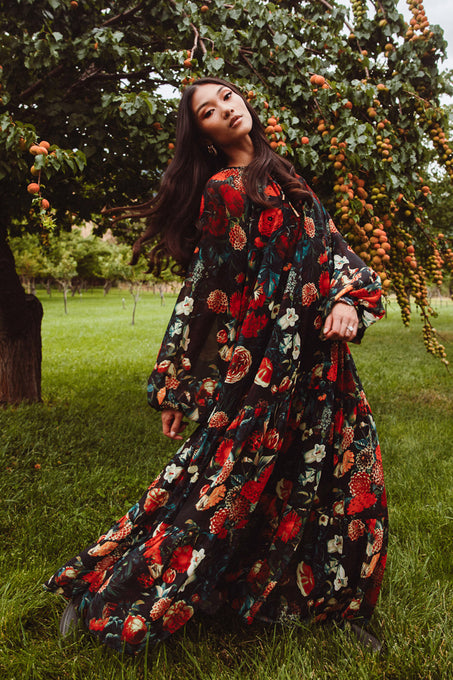A woman poses gracefully in a grassy orchard wearing the Ivy City Co Imogen Dress in Moody Floral—a flowing, elegant maxi dress that’s both maternity friendly and perfect for lush, nature-filled settings.