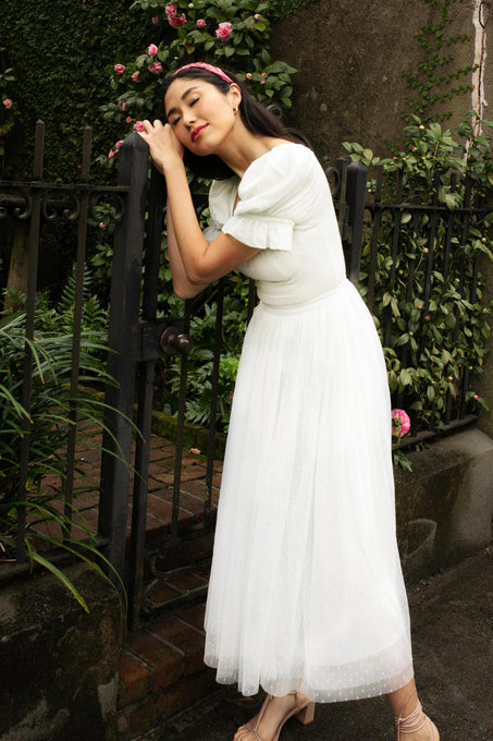 A woman wearing the Ivy City Co. Ballerina Dress in White leans against a black iron fence, eyes closed and smiling gently, surrounded by blooming pink roses and lush greenery.