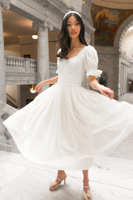 A woman twirls gracefully on marble steps inside a grand building, wearing the Ivy City Co. Ballerina Dress in White—a midi dress with puff sleeves and a full skirt.