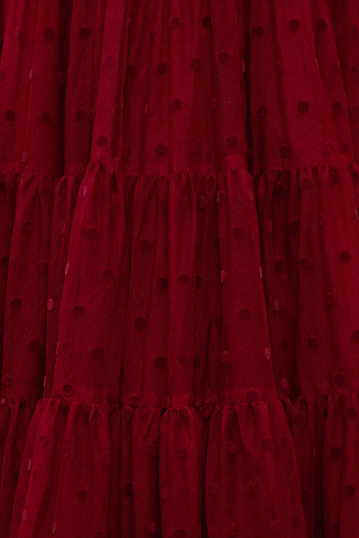 A close-up of the Solange Dress in Burgundy Tulle by Ivy City Co reveals deep red, sheer fabric adorned with a textured polka dot pattern and gathered, tiered sections that create a ruffled effect—an exquisite detail for an elegant or romantic holiday gown.