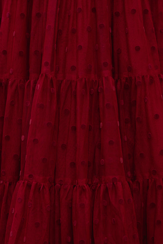 A close-up of the Solange Dress in Burgundy Tulle by Ivy City Co reveals deep red, sheer fabric adorned with a textured polka dot pattern and gathered, tiered sections that create a ruffled effect—an exquisite detail for an elegant or romantic holiday gown.