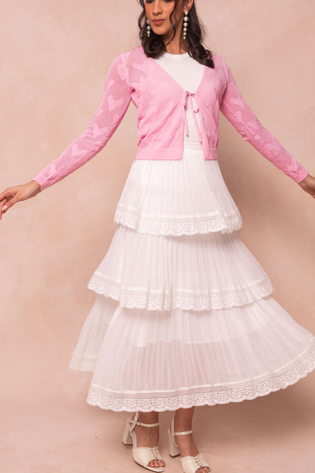 A woman wears the Ivy City Co With Love Sweater Cardigan in pink over a white top and a long tiered white lace-trim skirt with white open-toe heels, standing against a soft beige background.