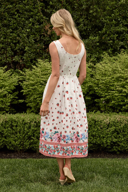 A woman stands outdoors on grass with green bushes in the background, wearing the Ivy City Co Sadie Dress in Strawberry Vintage Tier—a white cotton summer dress featuring red and pink floral patterns.