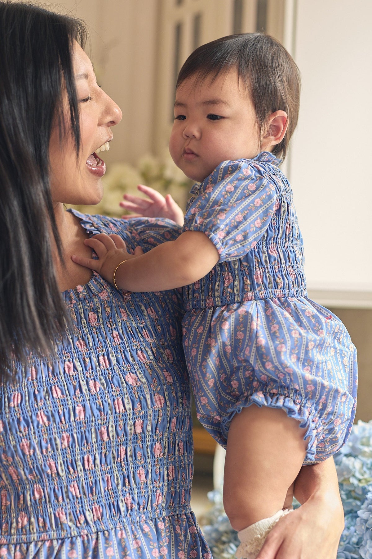 A woman holds a baby as they both wear matching blue patterned outfits. The woman smiles warmly at the baby, who looks off to the side in her adorable Ivy City Co Baby Mariah Romper in Linear Print—a cozy cotton snap romper featuring a playful floral-inspired pattern.