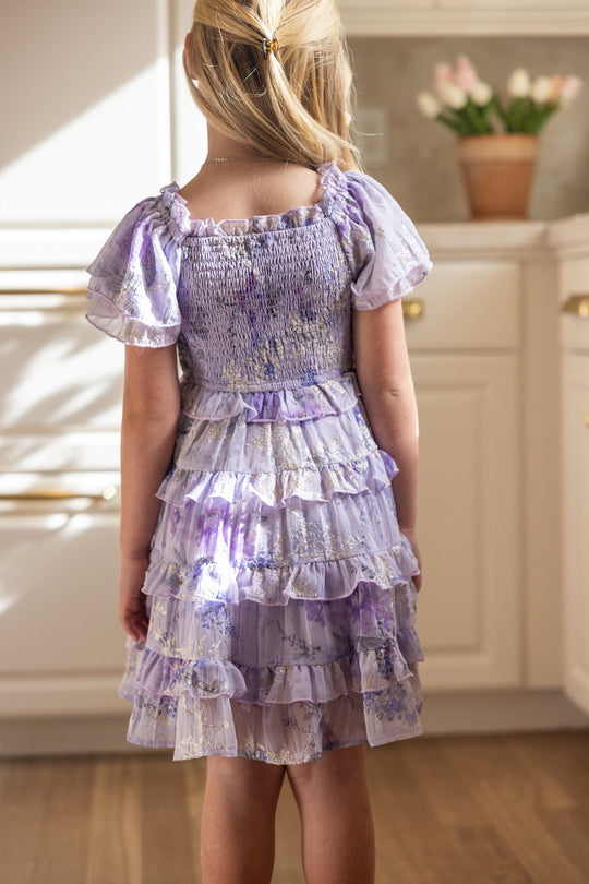 A young girl with blonde hair in a clip stands in a kitchen, wearing the Ivy City Co Mini Winnie Dress in Lavender Aquarelle Fleur—a ruffled, tiered dress adorned with a lovely purple floral print. She is facing away from the camera, while a pot of tulips sits on the counter in the background.