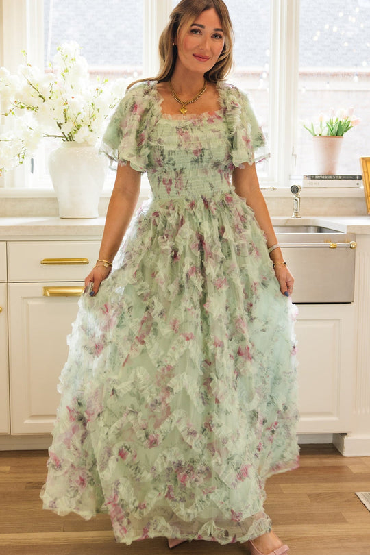 A woman in the Florentina Dress in Pistachio Aquarelle Fleur by Ivy City Co stands in a bright kitchen with white cabinets, a wood floor, and a large window. She smiles and gently holds out her nursing-friendly chiffon dress, which features a pastel green floral design. White flowers and various kitchen items are visible behind her, highlighting the comfort and style this dress offers for any occasion.