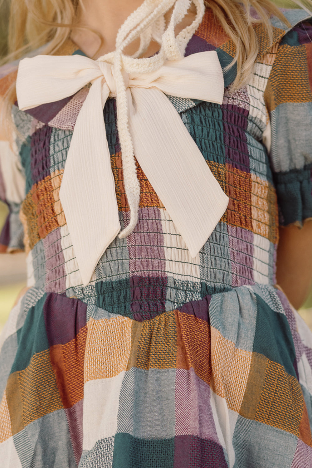 A close-up of a girl wearing the Ivy City Co Mini Alice Dress in Maize Gingham, featuring a large cream-colored bow at the collar, a smocked bodice, puff sleeves, and intricate detailing. Only her neck and part of her face are visible.
