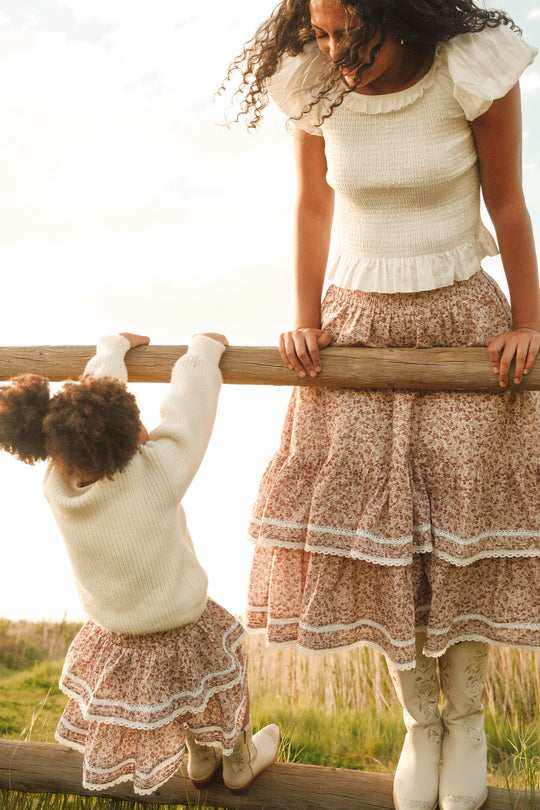 A woman and a young girl enjoy the outdoors in matching white sweaters and Ivy City Co's Mini Annalise Skirt in Philocaly Floral. The girl's knee-length floral skirt completes her look, while both wear white boots for a coordinated style.