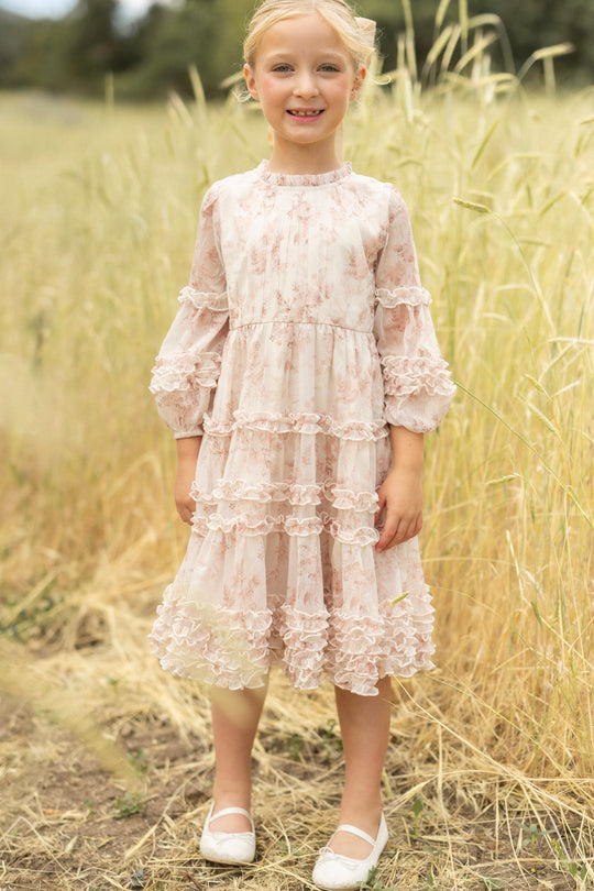 A young girl stands in a field, smiling and wearing the Ivy City Co Mini Geneva Dress in Banana Creme Aquarelle Fleur—a light yellow, ruffled flower girl dress with long sleeves and white flats. The background is softly blurred greenery.