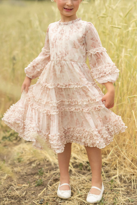 A young child stands in a grassy field, smiling and holding out the sides of her Ivy City Co Mini Geneva Dress in Banana Creme Aquarelle Fleur, a long-sleeved, ruffled floral dress. She pairs it with white shoes.