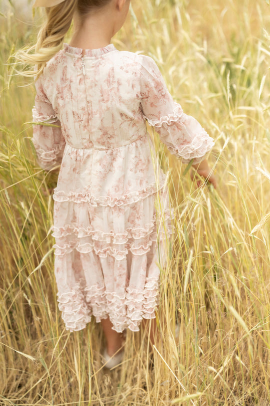 A young girl in the Mini Geneva Dress in Banana Creme Aquarelle Fleur by Ivy City Co stands in a field of tall, golden grass with her blonde hair tied back in a ponytail, facing away from the camera.
