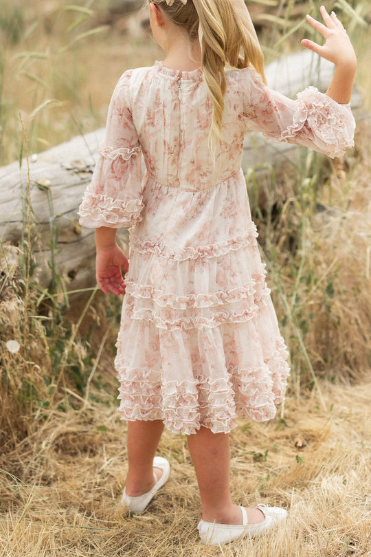 A young girl with blonde hair in a ponytail, wearing the Ivy City Co Mini Geneva Dress in Banana Creme Aquarelle Fleur and white shoes, stands outdoors on dry grass, facing away as she waves beside a weathered log.