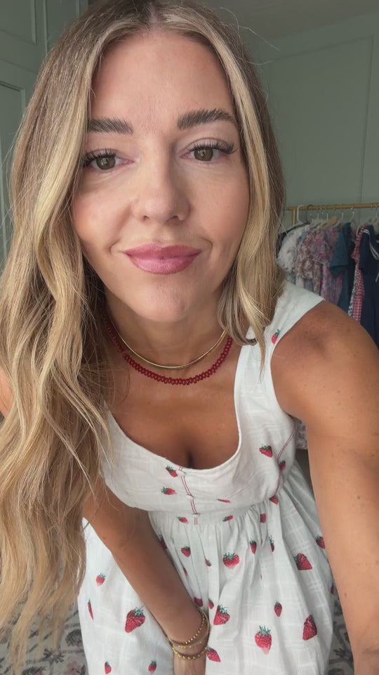 A woman with long blonde hair wearing the Ivy City Co Sadie Dress in Strawberry Vintage Tier and a red beaded necklace smiles at the camera, with clothes hanging on a rack in the background.