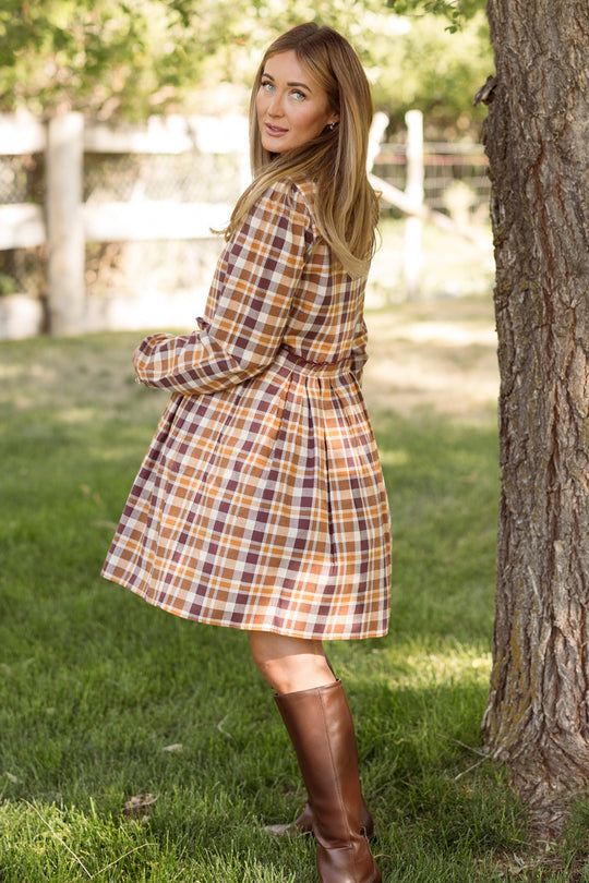 A woman in the Ivy City Co Bowie Long Sleeve Dress in Spiced Plaid, paired with tall brown boots, stands on grass near a tree, looking back over her shoulder.