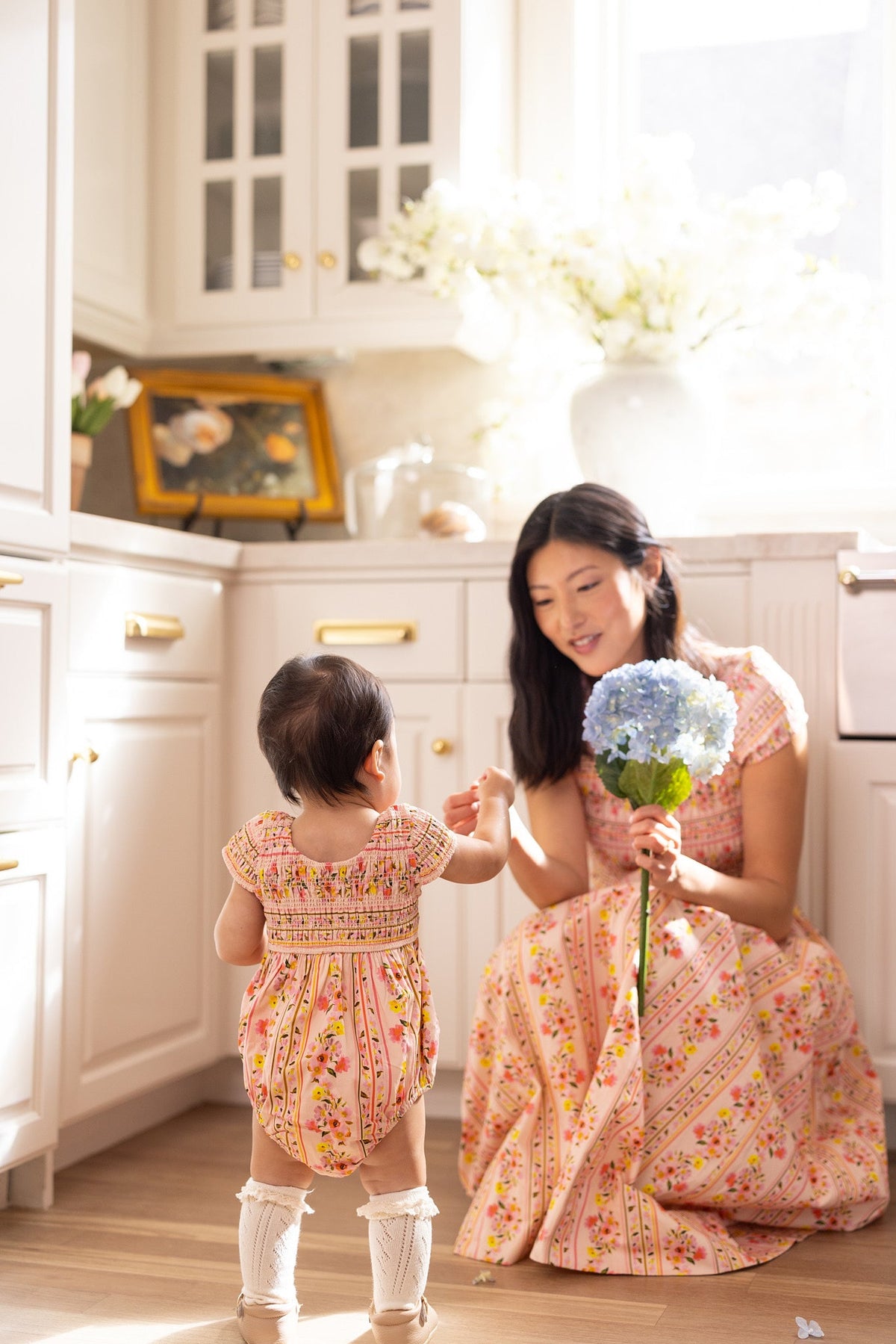In a sunlit kitchen, a woman in a floral dress kneels, holding blue hydrangeas. A toddler wearing the Ivy City Co Baby Marlow Romper in Peach Handpainted Flora Stripe stands facing her and reaches out as sunlight streams through the window.