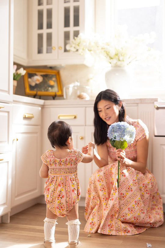 In a sunlit kitchen, a woman in a floral dress kneels, holding blue hydrangeas. A toddler wearing the Ivy City Co Baby Marlow Romper in Peach Handpainted Flora Stripe stands facing her and reaches out as sunlight streams through the window.