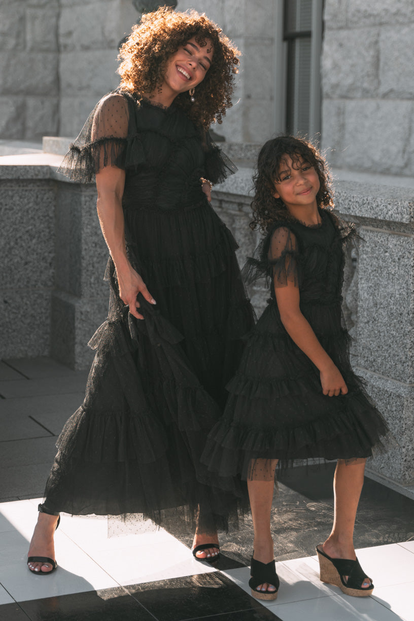 A woman and a young girl smile outdoors, both wearing matching Mini Whimsical Dresses in Black from Ivy City Co. Their layered tulle skirts and open-toed heels catch the sunlight as they stand close together by a stone wall, radiating joy.