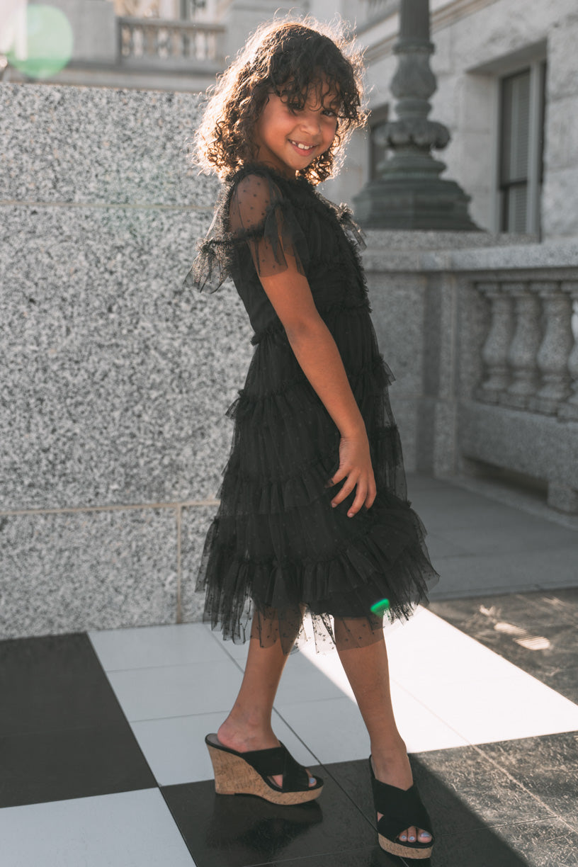 A young girl with curly hair smiles as she poses outdoors in the Mini Whimsical Dress in Black by Ivy City Co, featuring a layered tulle skirt and wedge sandals, standing on a black-and-white tiled floor near a stone wall and railing.