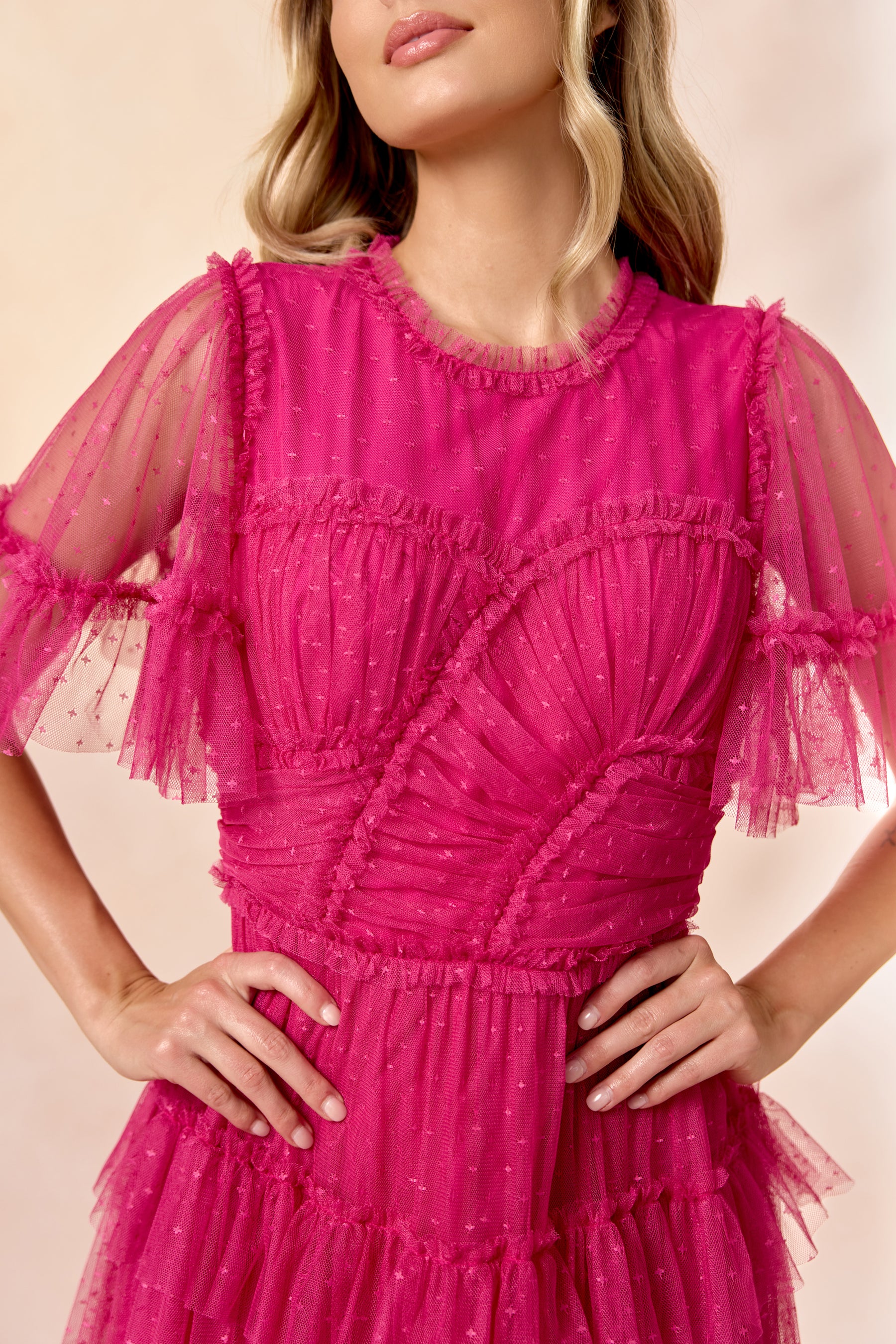 A woman models the Whimsical Dress in Fuchsia by Ivy City Co, featuring a ruffled tulle skirt and sheer dotted sleeves and bodice. She stands with hands on hips against a soft beige background, with only her upper body and part of her face visible.