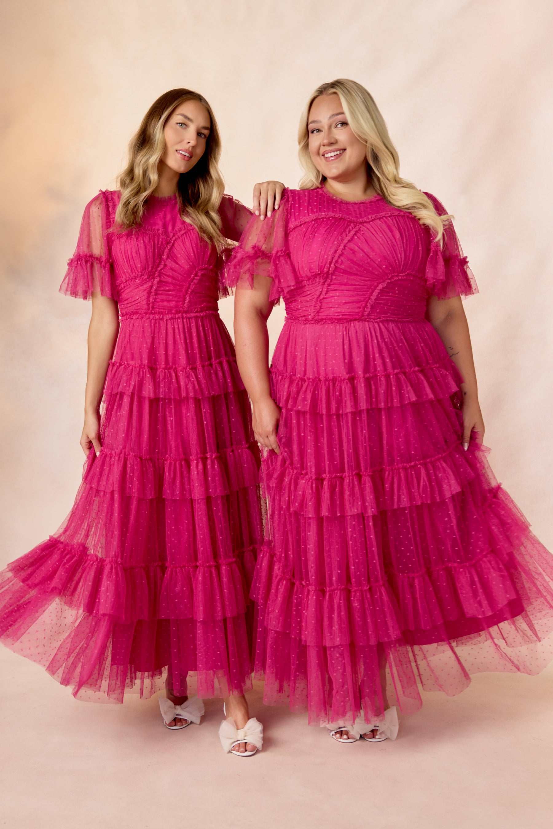 Two women smile side by side, wearing matching Whimsical Dress in Fuchsia by Ivy City Co, featuring ruffled tiered tulle skirts and sheer short sleeves, paired with white shoes against a soft, light background.
