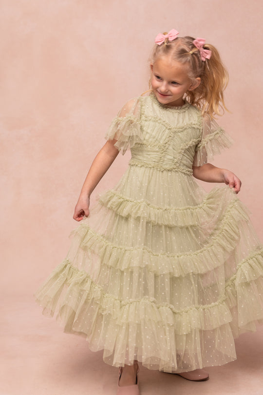 A young girl with blonde hair in pink bows smiles as she poses in the Ivy City Co Mini Whimsical Maxi Dress in Sage, featuring light green ruffles, sheer polka dot layers, and a twirl-worthy skirt against a soft beige background.