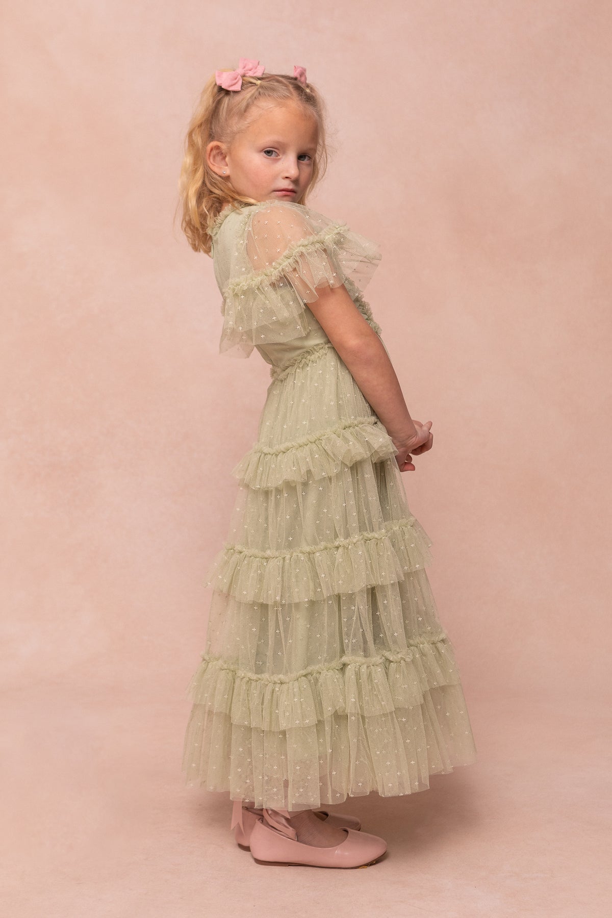 A young girl with blond hair and pink bows wears the Ivy City Co Mini Whimsical Maxi Dress in Sage, featuring flutter sleeves and a twirl-worthy skirt, styled with matching pink shoes against a light pink backdrop.