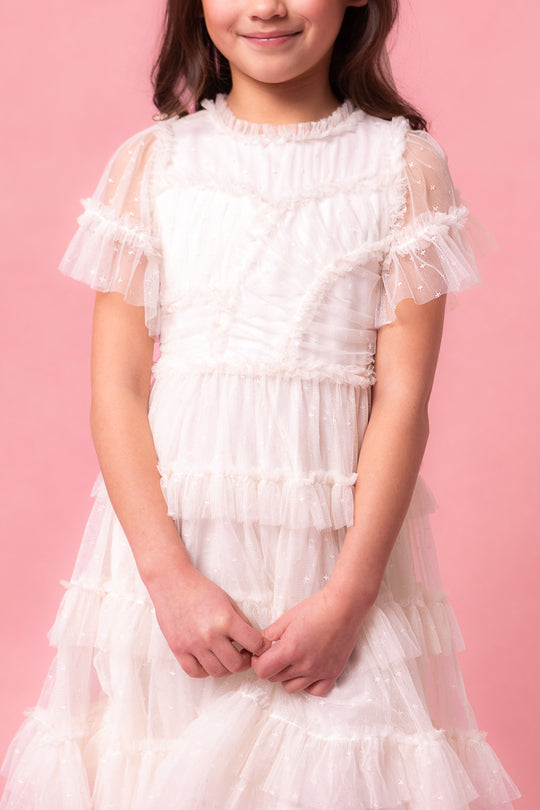 A young girl smiles softly, standing against a pink background in the Ivy City Co. Mini Whimsical Dress in White, featuring flutter sleeves and a layered tulle skirt with her hands clasped in front of her.