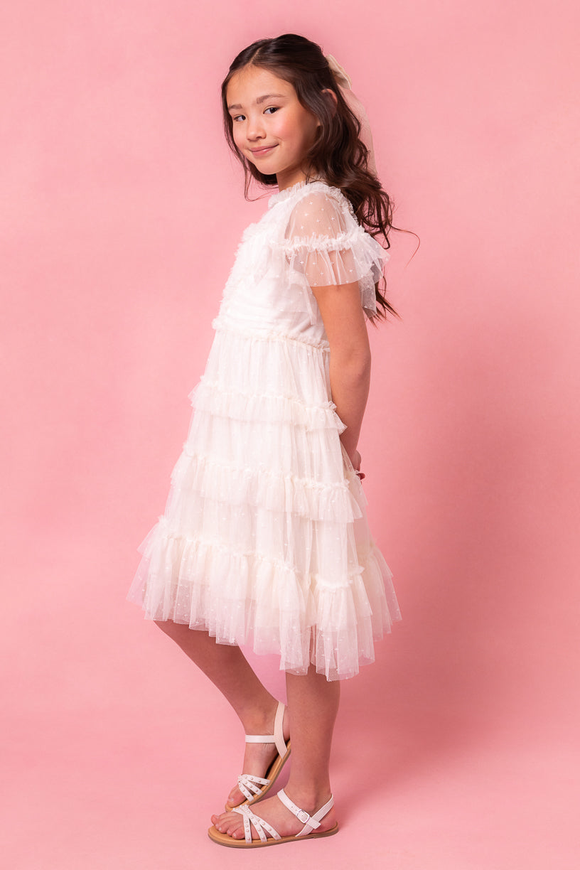 Young girl with long dark hair wearing the Ivy City Co Mini Whimsical Dress in White with flutter sleeves and a layered tulle skirt, paired with white sandals, standing sideways and smiling against a pink background.