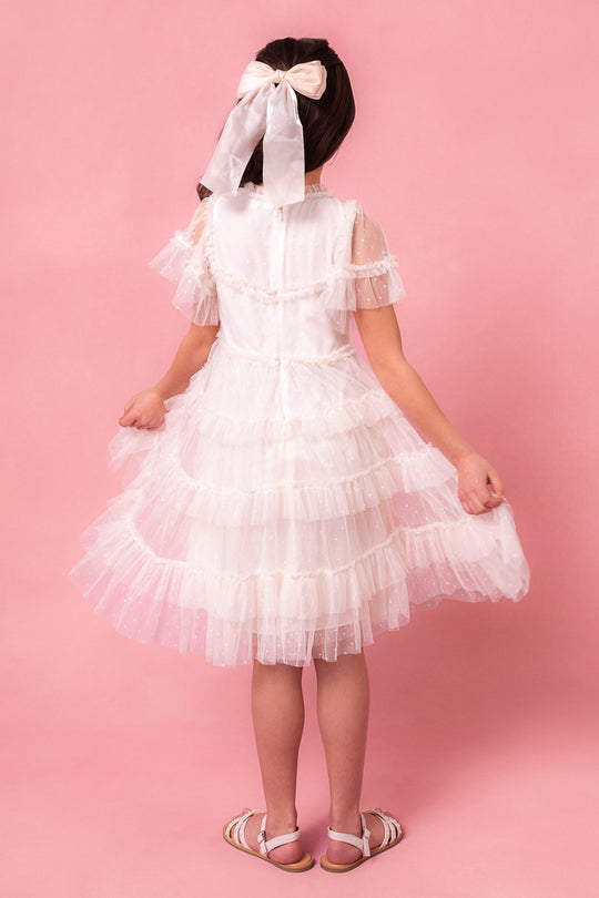 A young girl faces away against a pink background, wearing the Ivy City Co. Mini Whimsical Dress in White with flutter sleeves and a large white bow in her hair. She holds out the ruffled dress sides and wears matching white sandals.