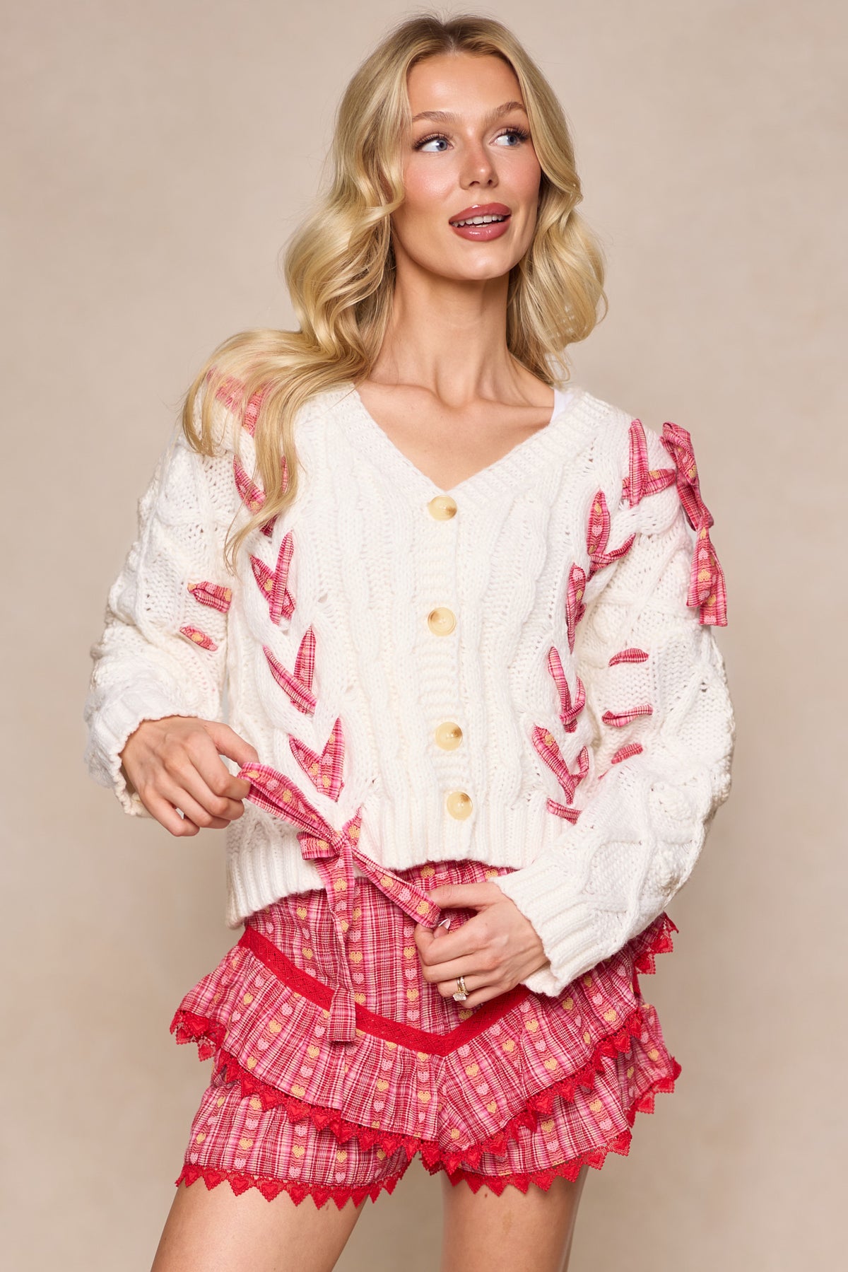 A woman with long blonde hair smiles while wearing the Ivy City Co Agatha Sweater in Ivory—an airy white knit cardigan with pink bows—paired with matching pink ruffled plaid shorts, set against a beige background.