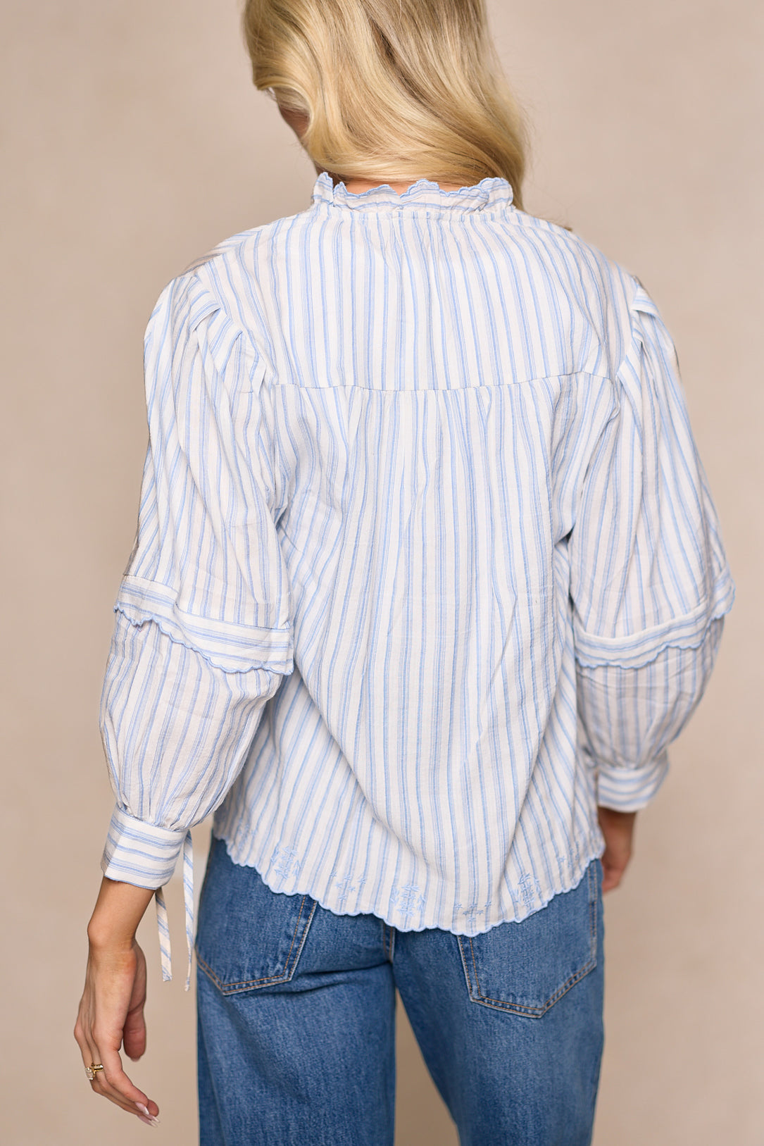 A woman with long blonde hair, seen from behind, wears the Ivy City Co Leticia Top in White & Blue Stripe—a cotton-poly blend with ruffle details and long sleeves—paired with blue jeans for a perfect spring look.