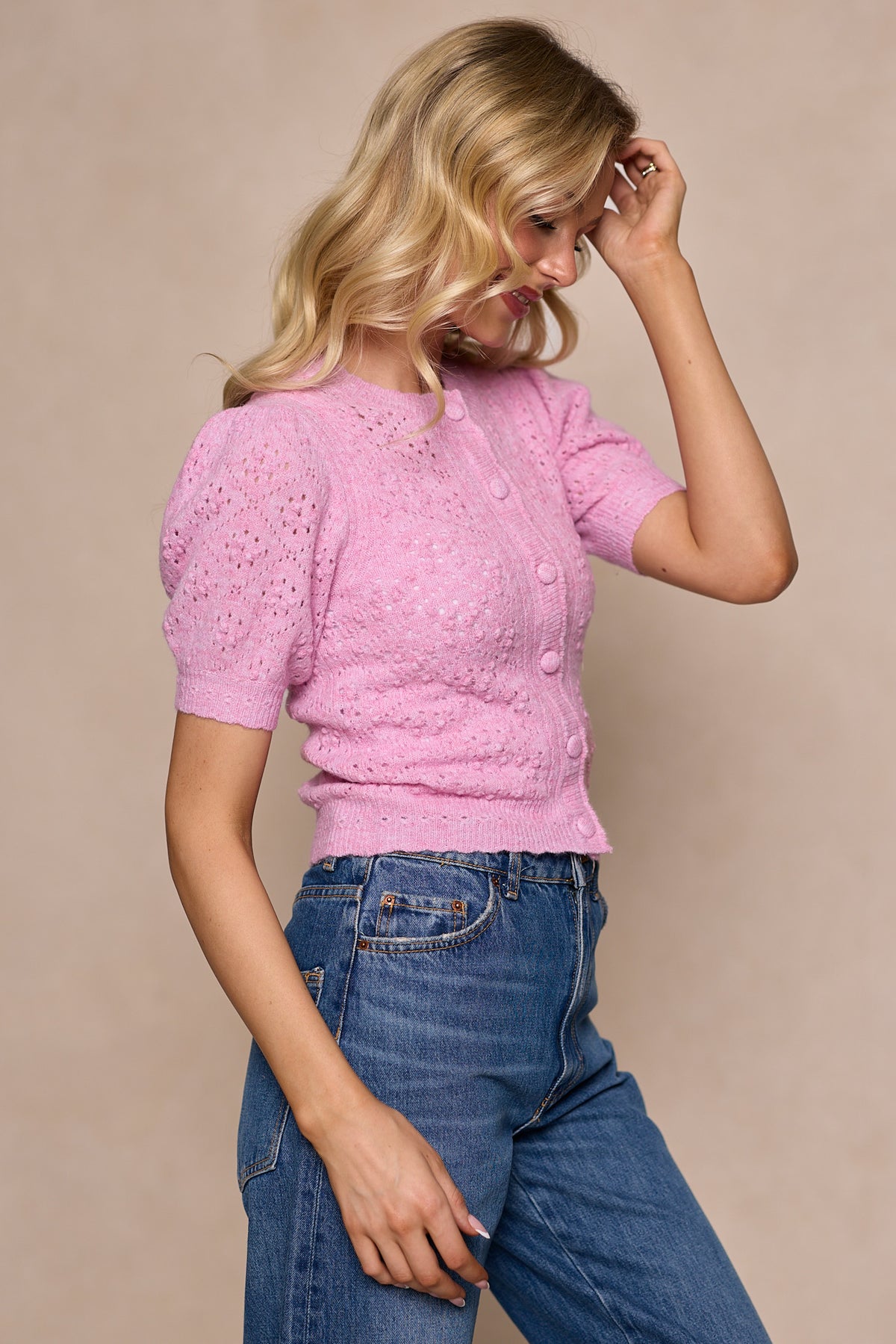 A woman with wavy blonde hair wears the Michaela Cardigan in Pink by Ivy City Co and high-waisted blue jeans. She stands sideways against a neutral background, touching her hair and looking down.