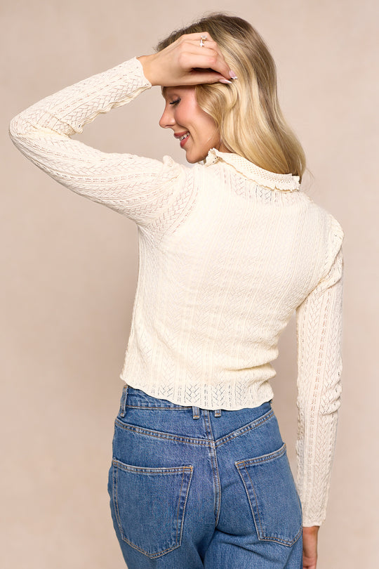 A woman with long blonde hair wears the Ivy City Co Odelia Cardigan in Cream and blue jeans, standing turned away while smiling and touching her head against a neutral background.