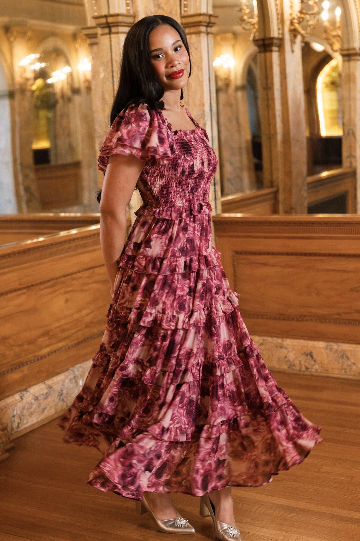 Clad in Ivy City Co’s Winnie Dress in Dreamy Roses—a flowing, ruffled pink floral holiday dress accented with delicate gold metallic stripes—a woman stands on polished wooden floors, smiling. Elegant arches, ornate mirrors, and warm lighting surround her, evoking a luxurious and classic atmosphere.