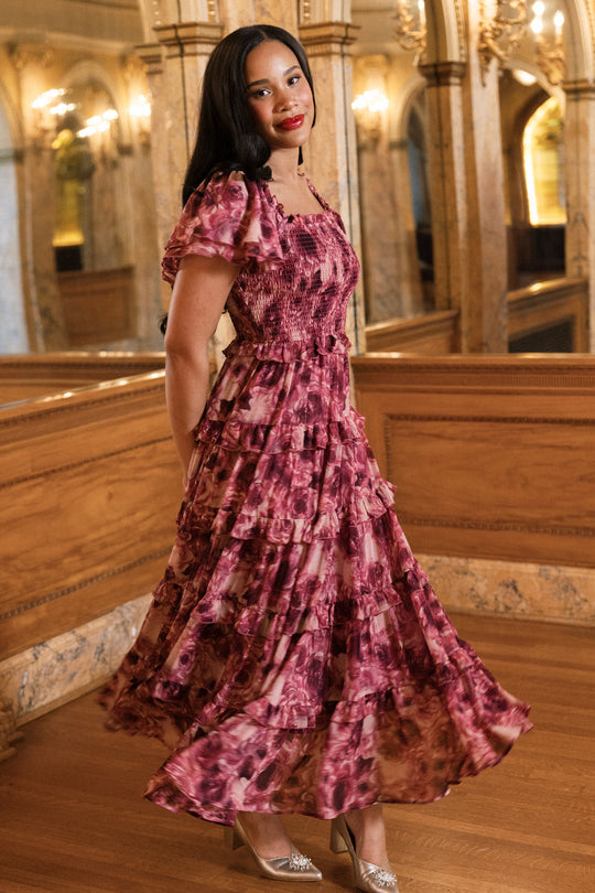 Clad in Ivy City Co’s Winnie Dress in Dreamy Roses—a flowing, ruffled pink floral holiday dress accented with delicate gold metallic stripes—a woman stands on polished wooden floors, smiling. Elegant arches, ornate mirrors, and warm lighting surround her, evoking a luxurious and classic atmosphere.